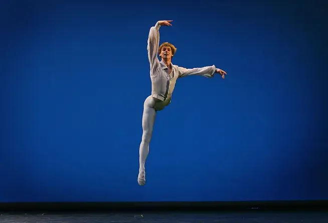 A ballet dancer in a light gray leotard and tights is captured mid-air in a graceful, extended pose against a solid blue…