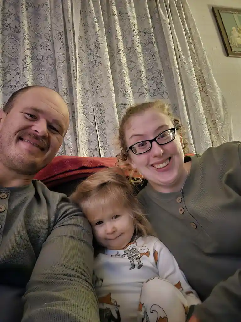 A selfie of a family of three — a man, a woman, and a young child — sitting together on a couch. The man and woman are…