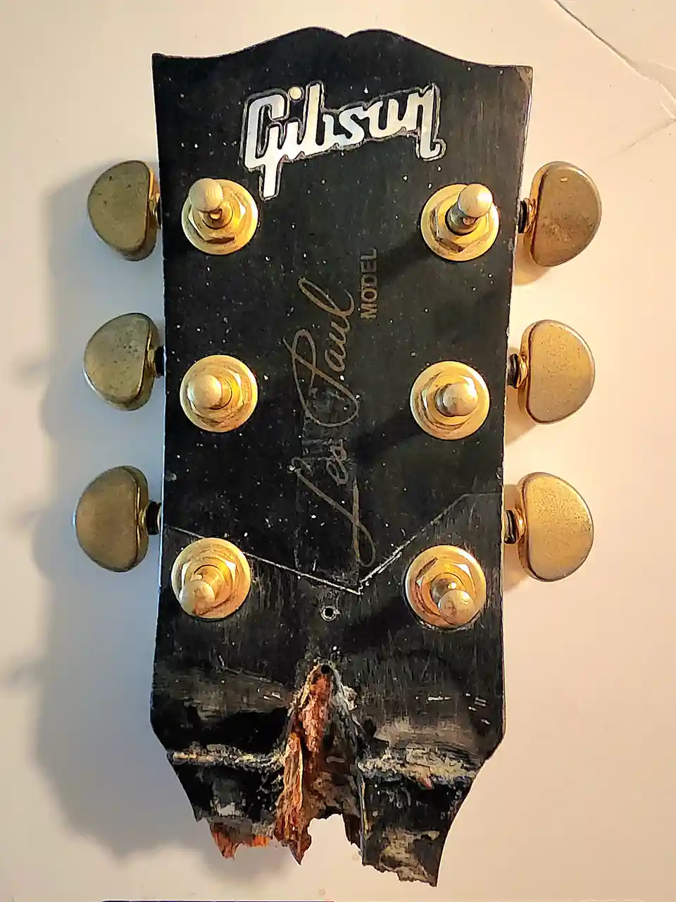 A close-up of a Gibson Les Paul guitar headstock with a heavily damaged, broken lower edge, revealing the wood underneath…