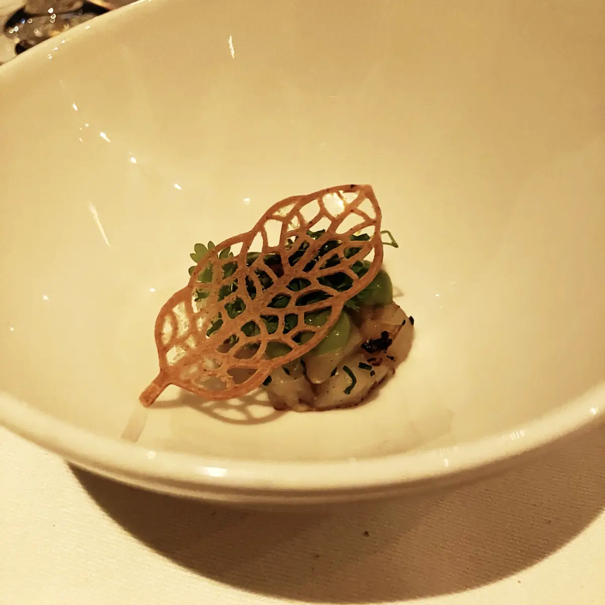 a close-up of a small, white ceramic bowl containing a tiny gourmet dish with green vegetables and a delicate, crispy, lacy…