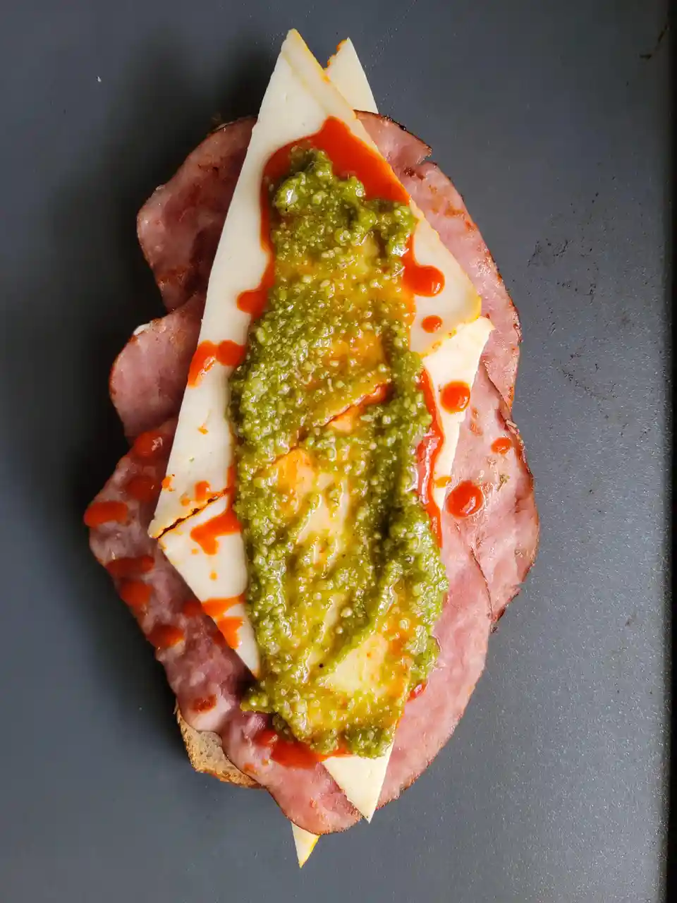 An overhead photo shows a sandwich on dark bread with layers of pink ham, white cheese slices, a thick green pesto sauce…