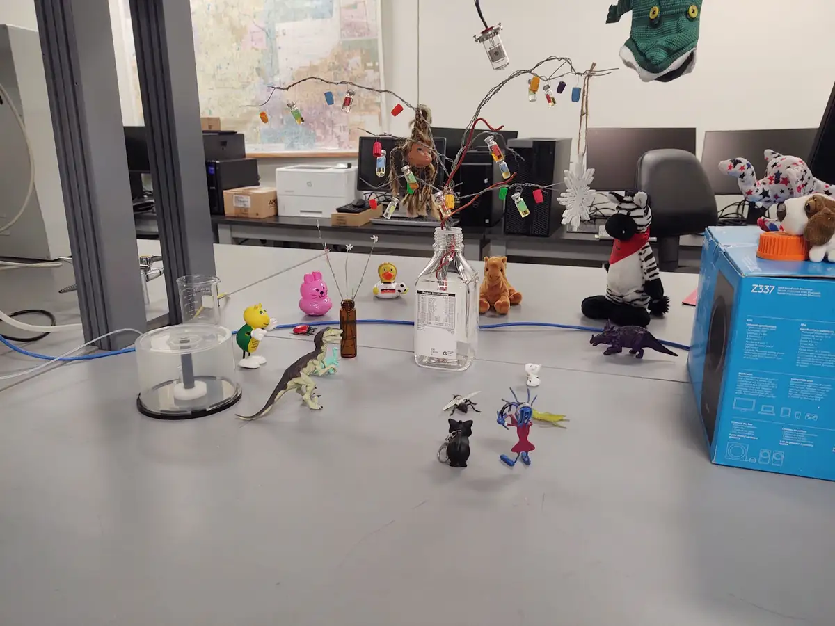 A cluttered office desk is covered with various toys, figurines, and tech items including a glass bottle with wires and tiny…