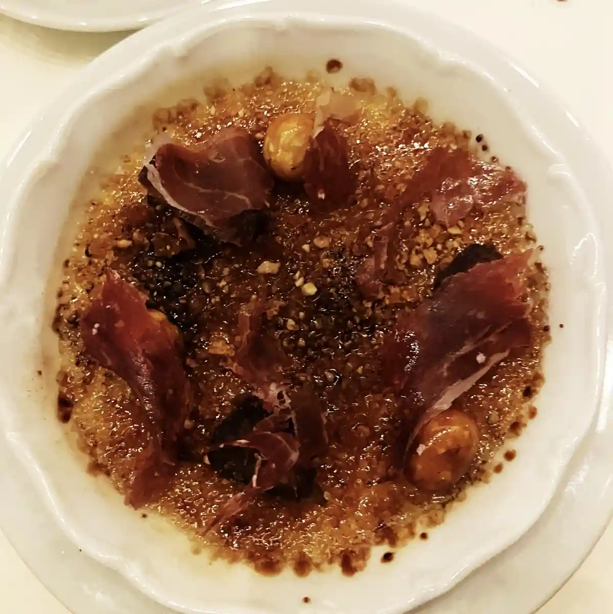 A close-up photo shows a white ceramic dish containing a dessert with a caramelized sugar crust, topped with thin slices of…