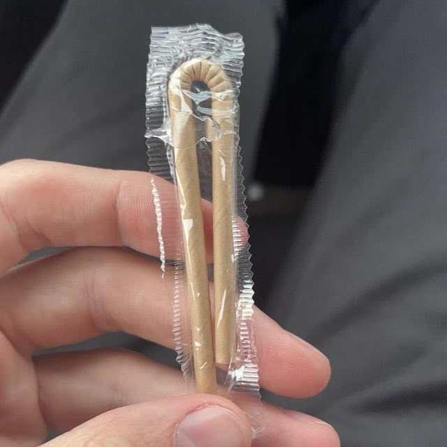 A hand holds a single, individually wrapped paper straw in clear plastic packaging. The straw is bent into a loop shape and…