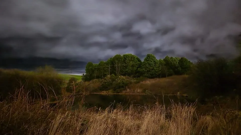 A moody landscape photo shows a dark, stormy sky looming over a calm pond surrounded by trees and dry grass. The overlaid…