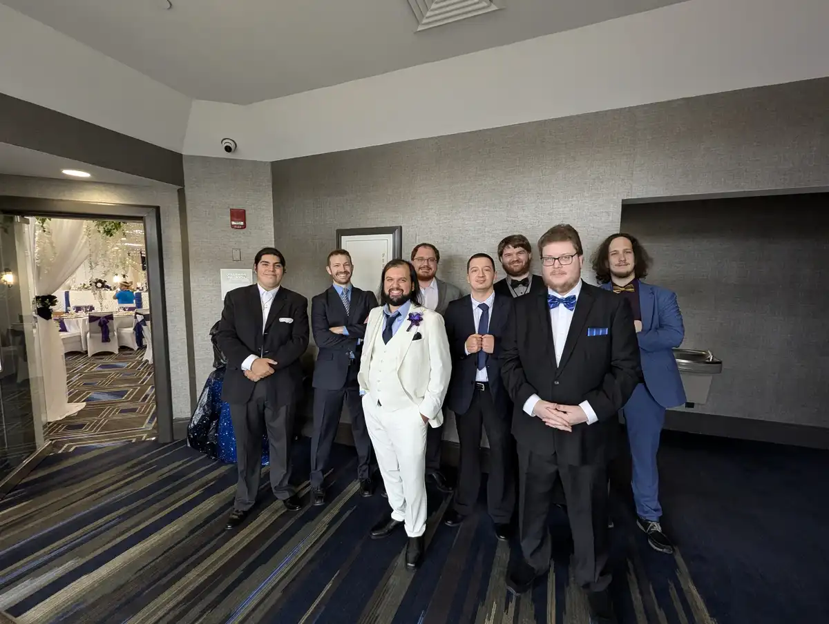 A group of eight men in formal suits pose for a photo in a hotel hallway, with a wedding reception visible through a doorway…