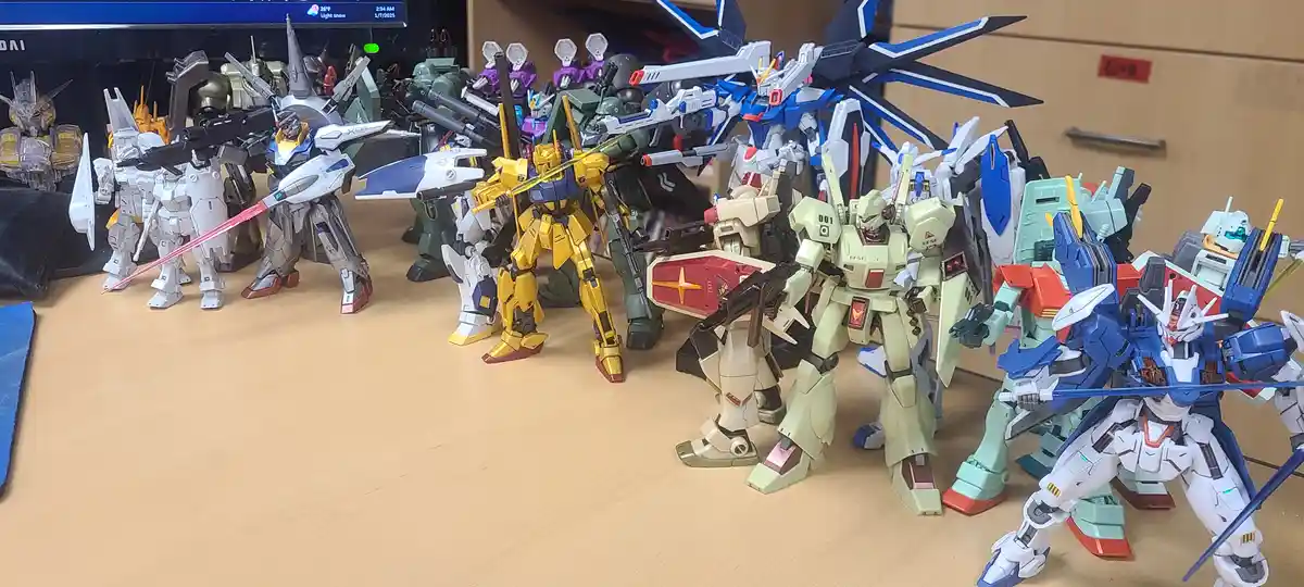 A wide shot shows dozens of colorful, detailed gundam model kits arranged in rows on a wooden desk, with a computer monitor…