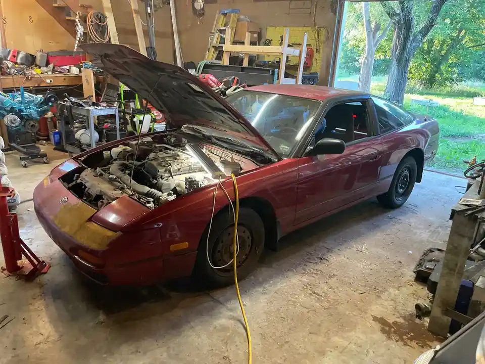 A maroon Nissan 300ZX is parked in a cluttered garage with its hood open, revealing the engine bay. A yellow work light is…