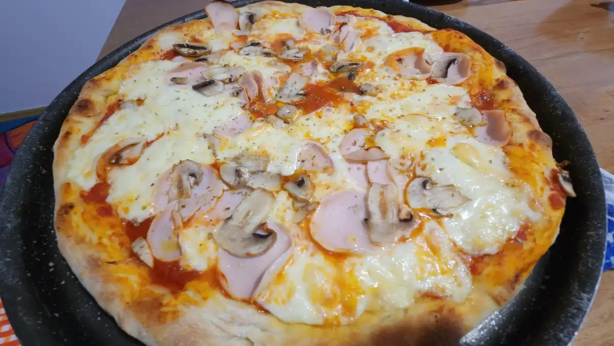 A freshly baked pizza with melted cheese, mushrooms, and ham slices sits in a black pan on a wooden table. The text overlay…