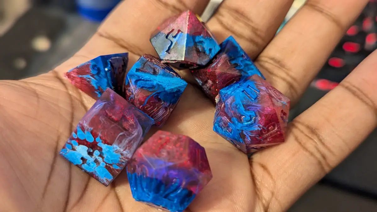 A hand holds a collection of custom-made, colorful polyhedral dice with swirling blue and red resin and glitter, typical of…