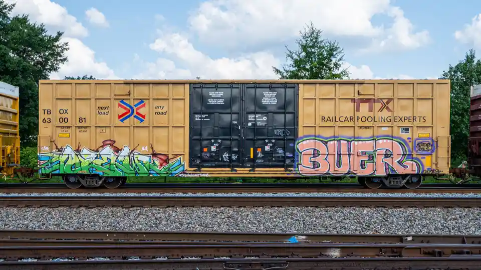 A yellow railcar with graffiti, including the word 'BUER' in large pink letters, sits on train tracks under a partly cloudy…