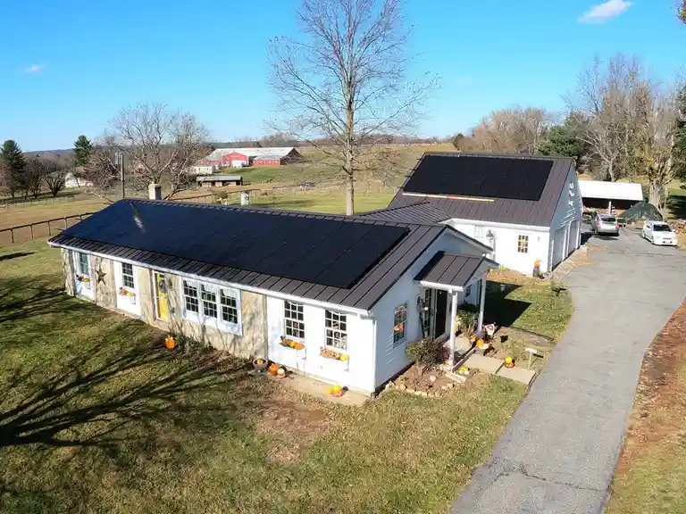 An aerial photo shows two white farmhouses with dark metal roofs, both covered in large black solar panels. The houses are…