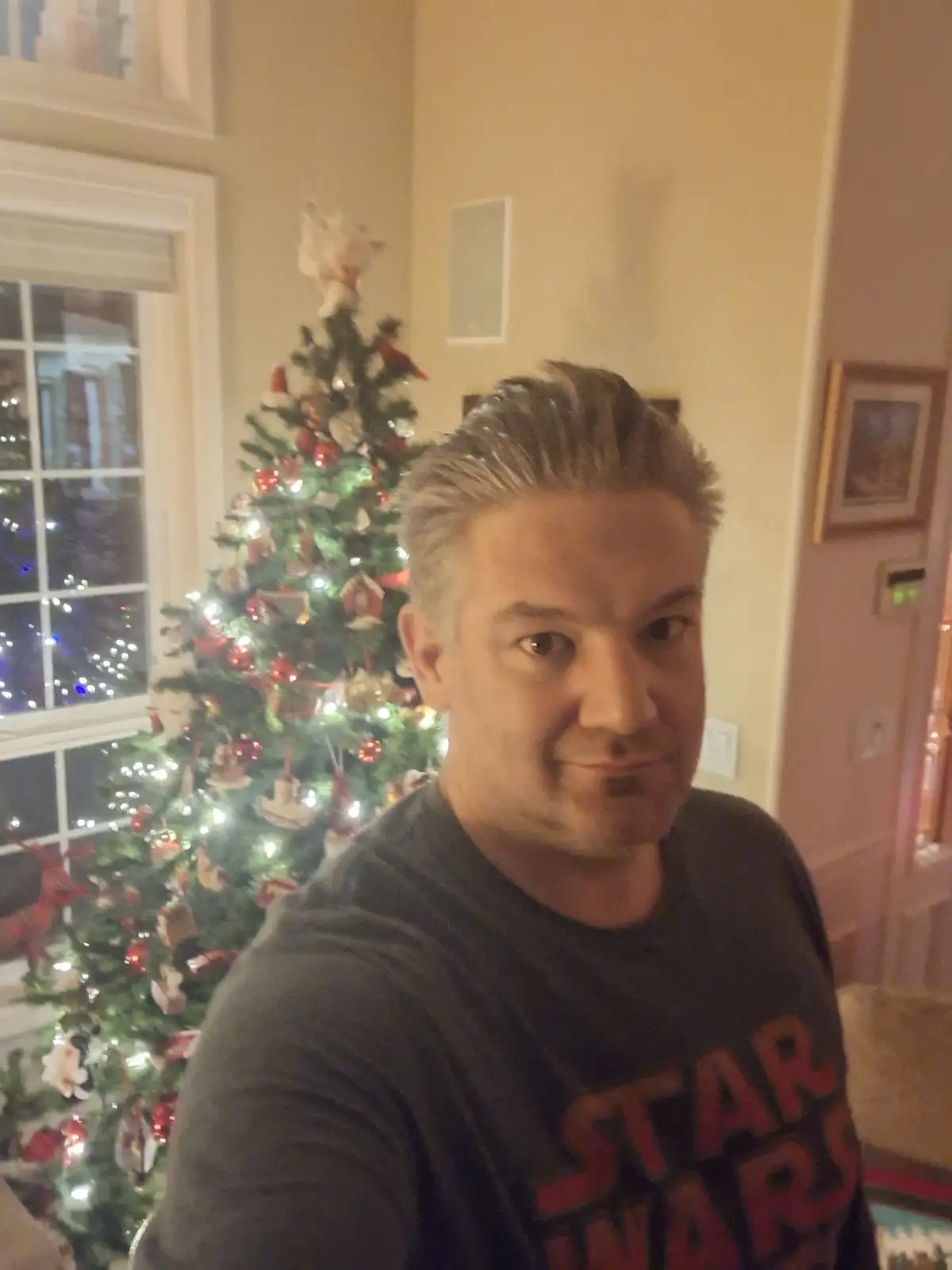 A man with styled grayish hair and a slight smirk wears a dark t-shirt with 'STAR WARS' in red letters, standing indoors in…