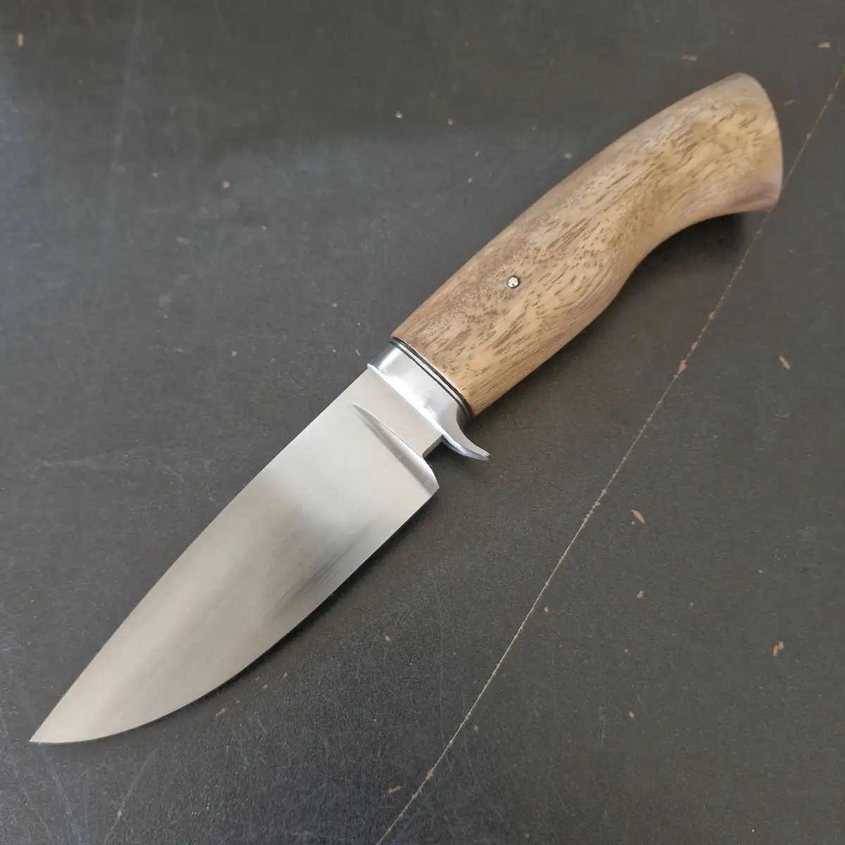A close-up photo shows a handmade knife with a wooden handle and a brushed metal blade resting on a dark, scratched surface…