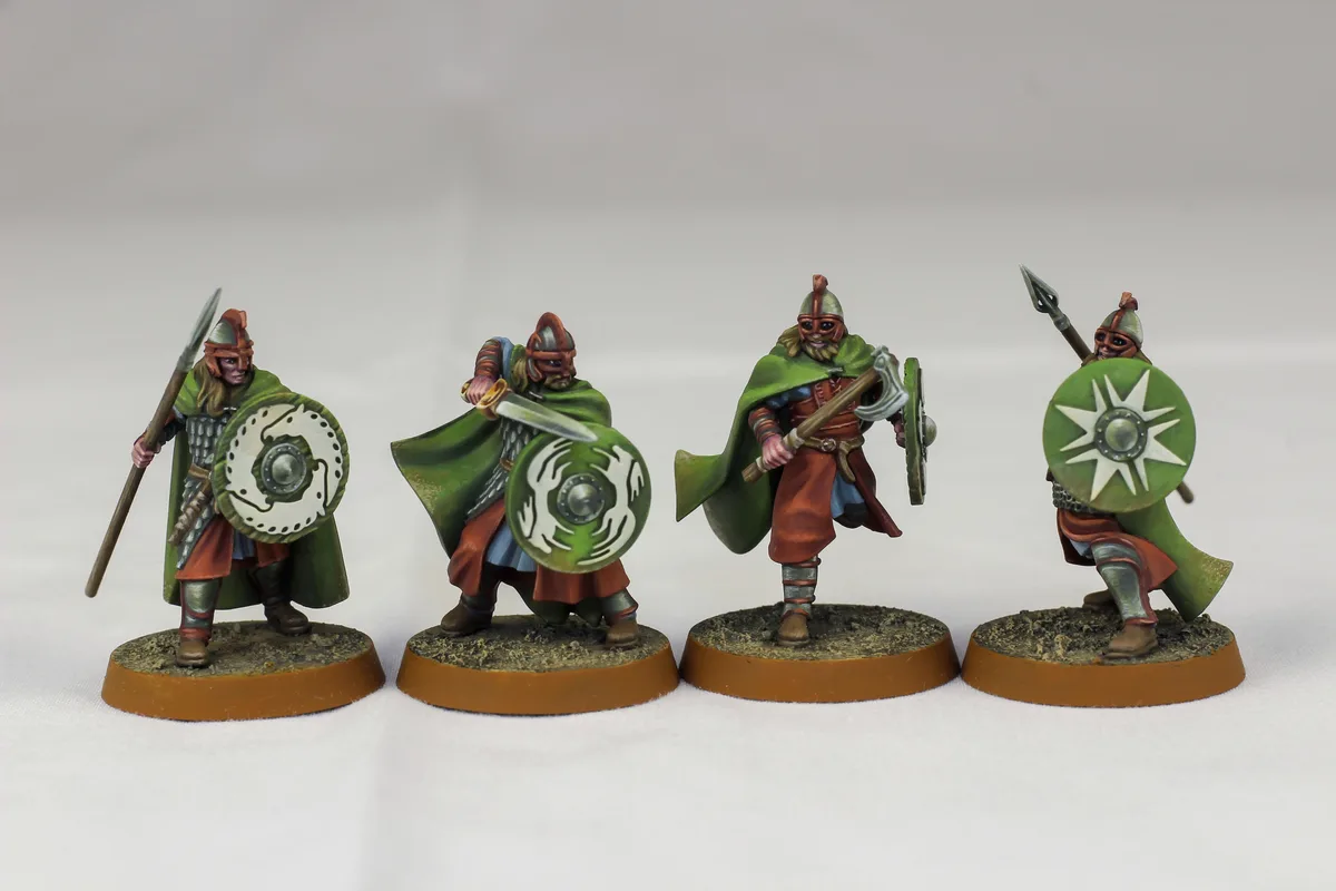 Four painted miniature fantasy warriors in green cloaks and armor stand in a row on circular bases against a white…