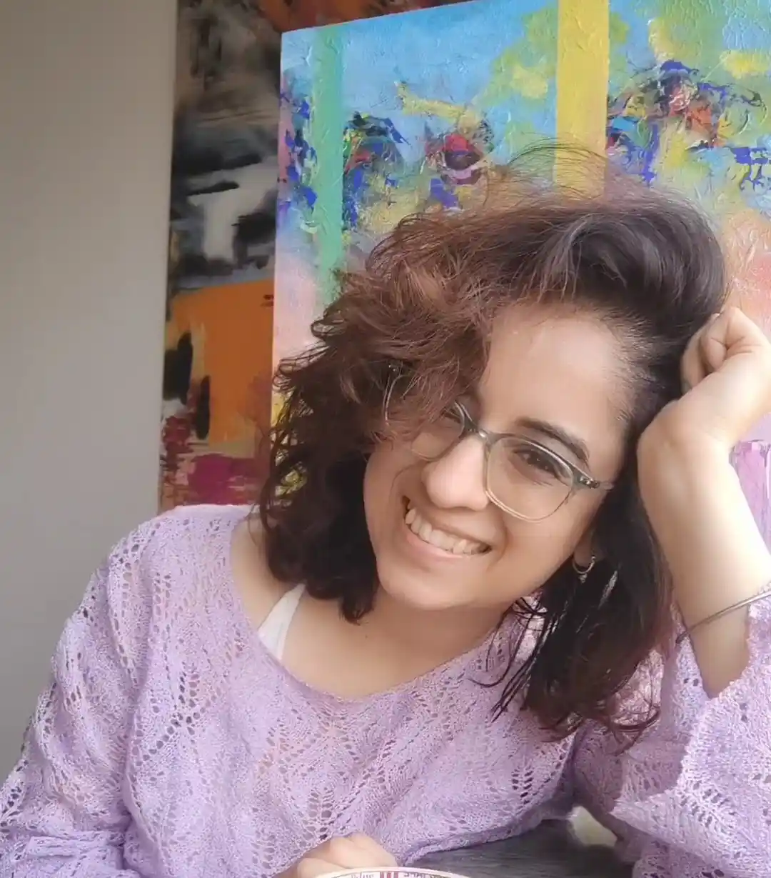 a woman with curly brown hair and glasses smiles while resting her head on her hand, wearing a purple lace top, with a…