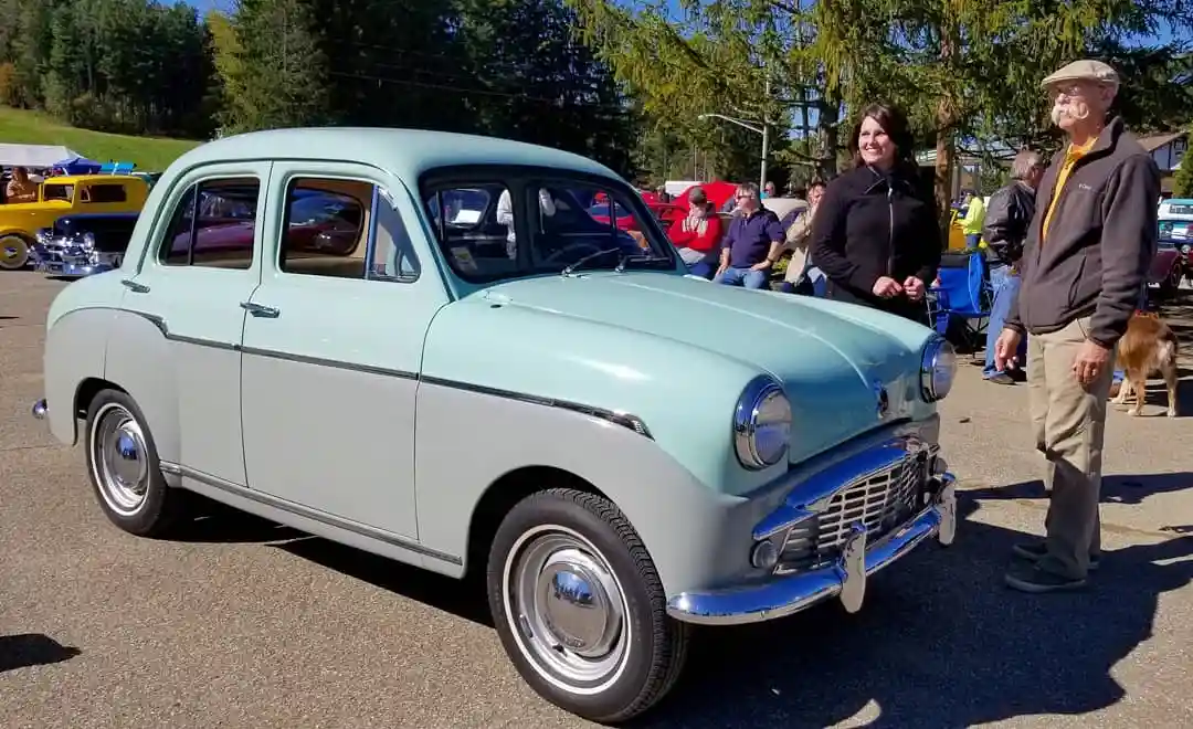 A vintage two-tone light blue and silver car is parked outdoors at what appears to be a car show, with people milling about…