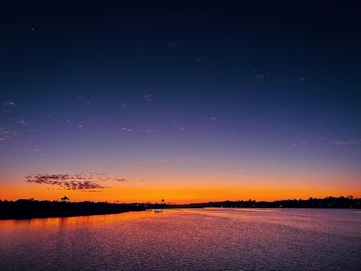 A serene twilight photo shows a calm body of water reflecting vibrant orange and purple hues from the sky, with a distant…