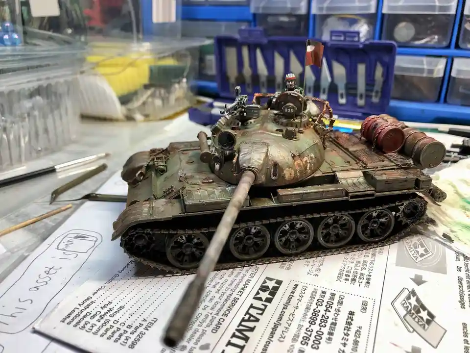 A highly detailed, weathered green tank model sits on a workbench surrounded by hobby tools and storage bins. The tank has a…