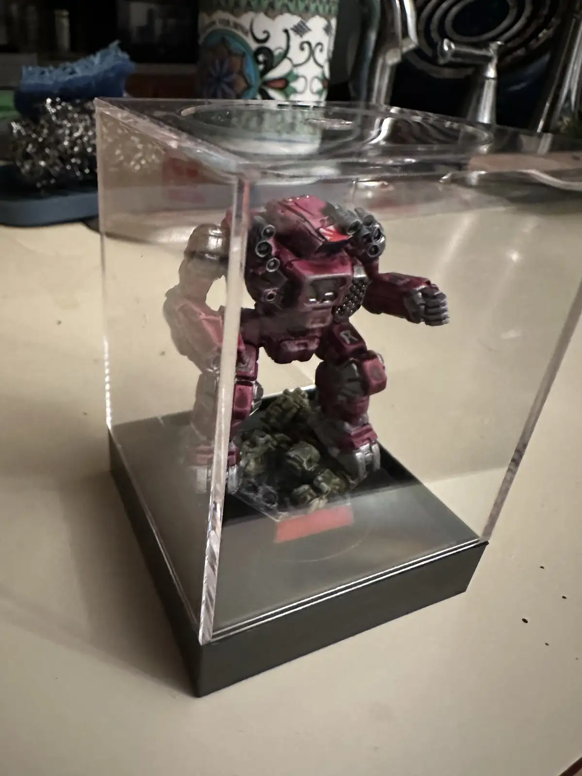 A red and grey mech figurine, resembling a Warhammer 40k Land Raider or similar sci-fi vehicle, is displayed inside a clear…