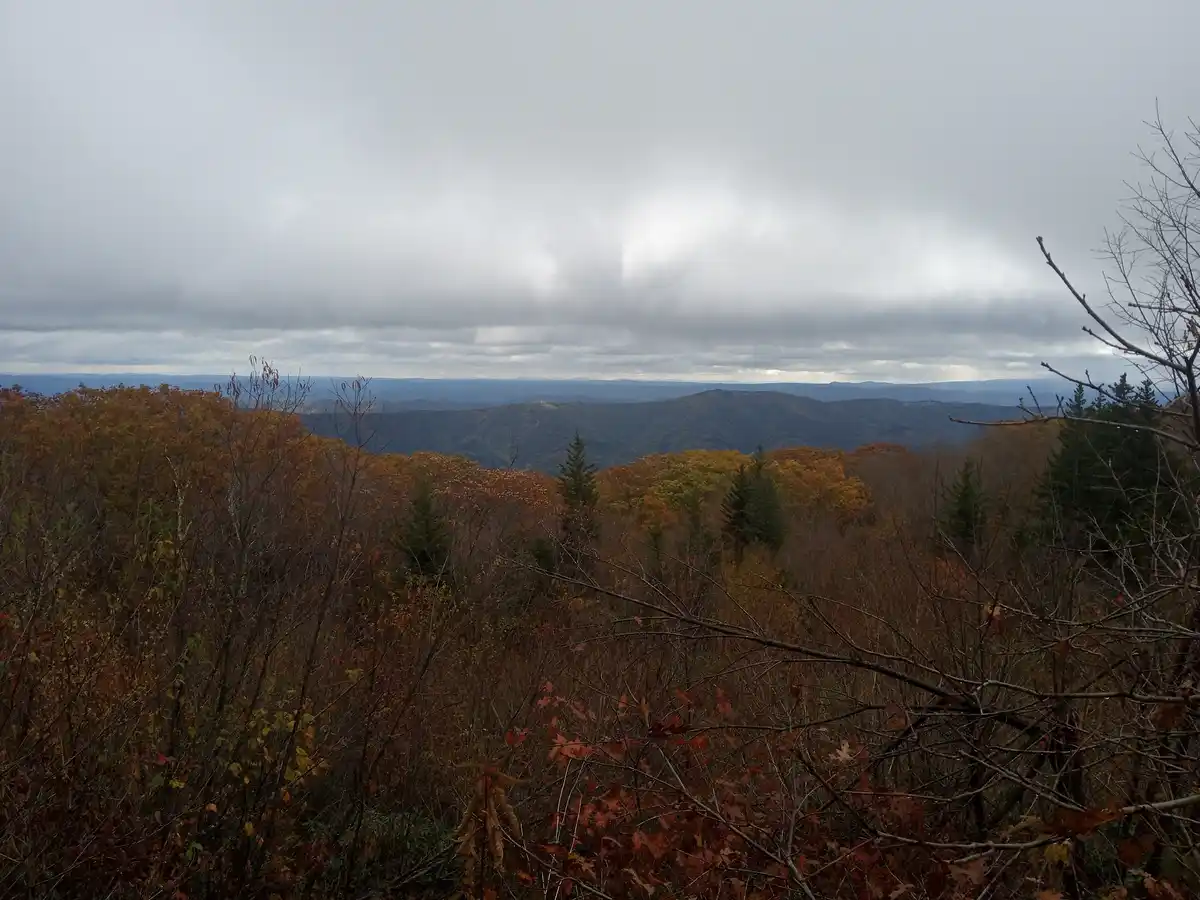 A wide-angle photo shows a cloudy, overcast sky above rolling mountain ranges covered in autumn foliage of orange, red, and…