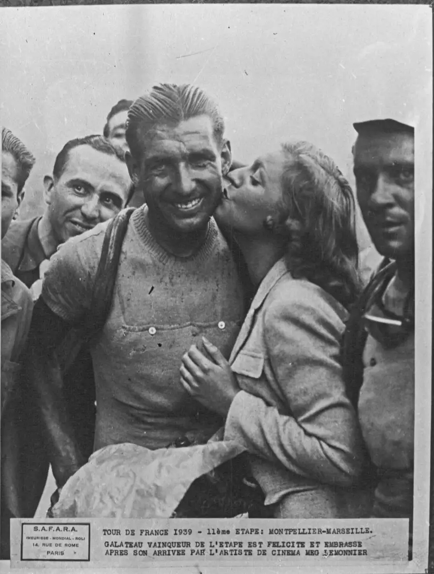 A vintage black-and-white photo from the 1939 Tour de France shows cyclist Galateau, winner of stage 11, smiling broadly as…