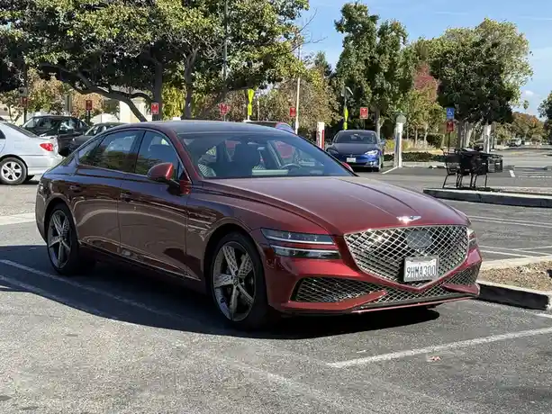 A maroon Genesis G80 sedan is parked in a sunny outdoor parking lot, with a blue Tesla visible in the background. The car's…