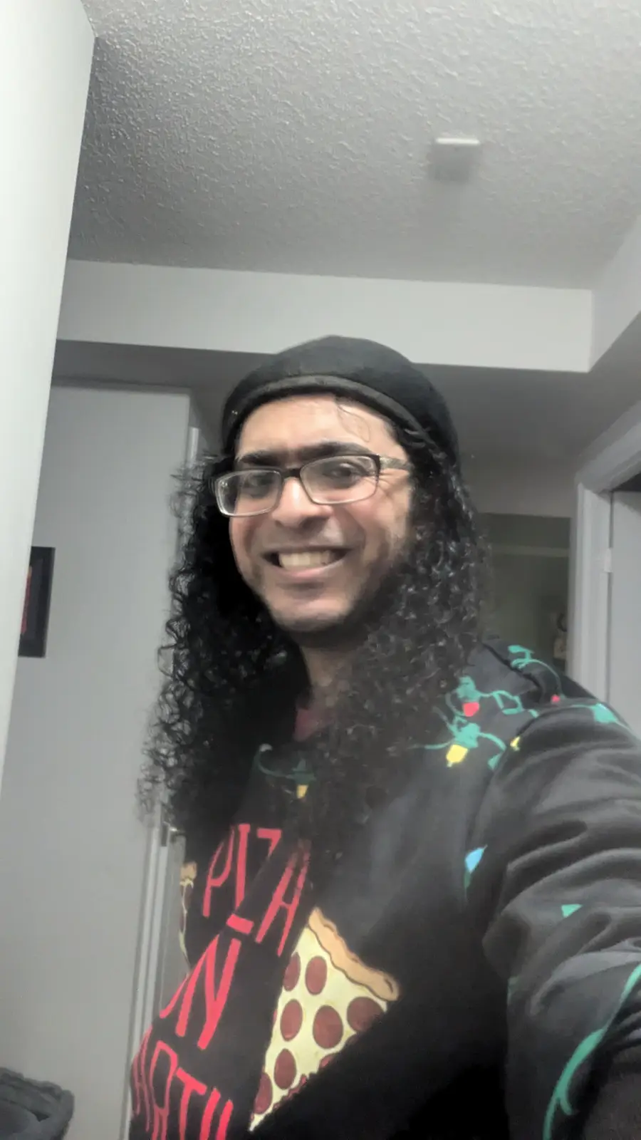 A person with long curly hair, glasses, and a black beret smiles broadly while taking a selfie indoors. They wear a black…