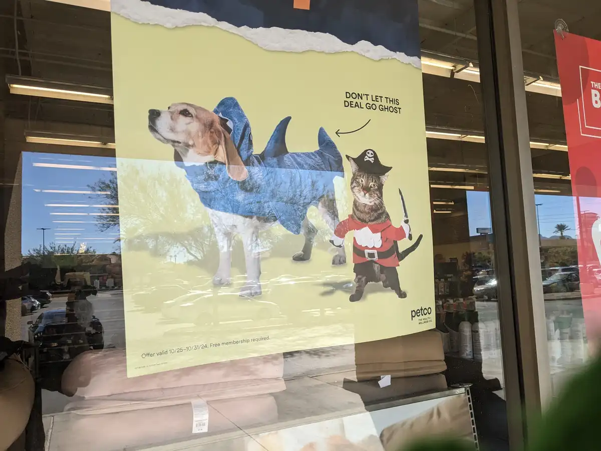 A Petco promotional poster taped to a store window shows a beagle in a blue shark costume and a tabby cat in a pirate…