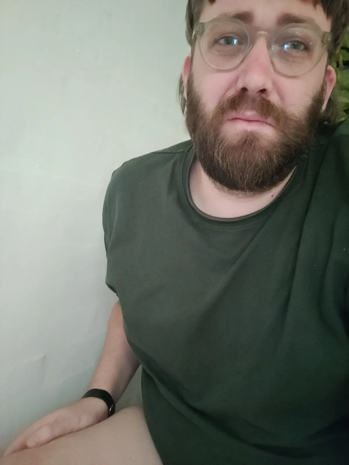 A man with a beard and glasses wearing a green t-shirt looks directly at the camera with a weary, resigned expression. He is…