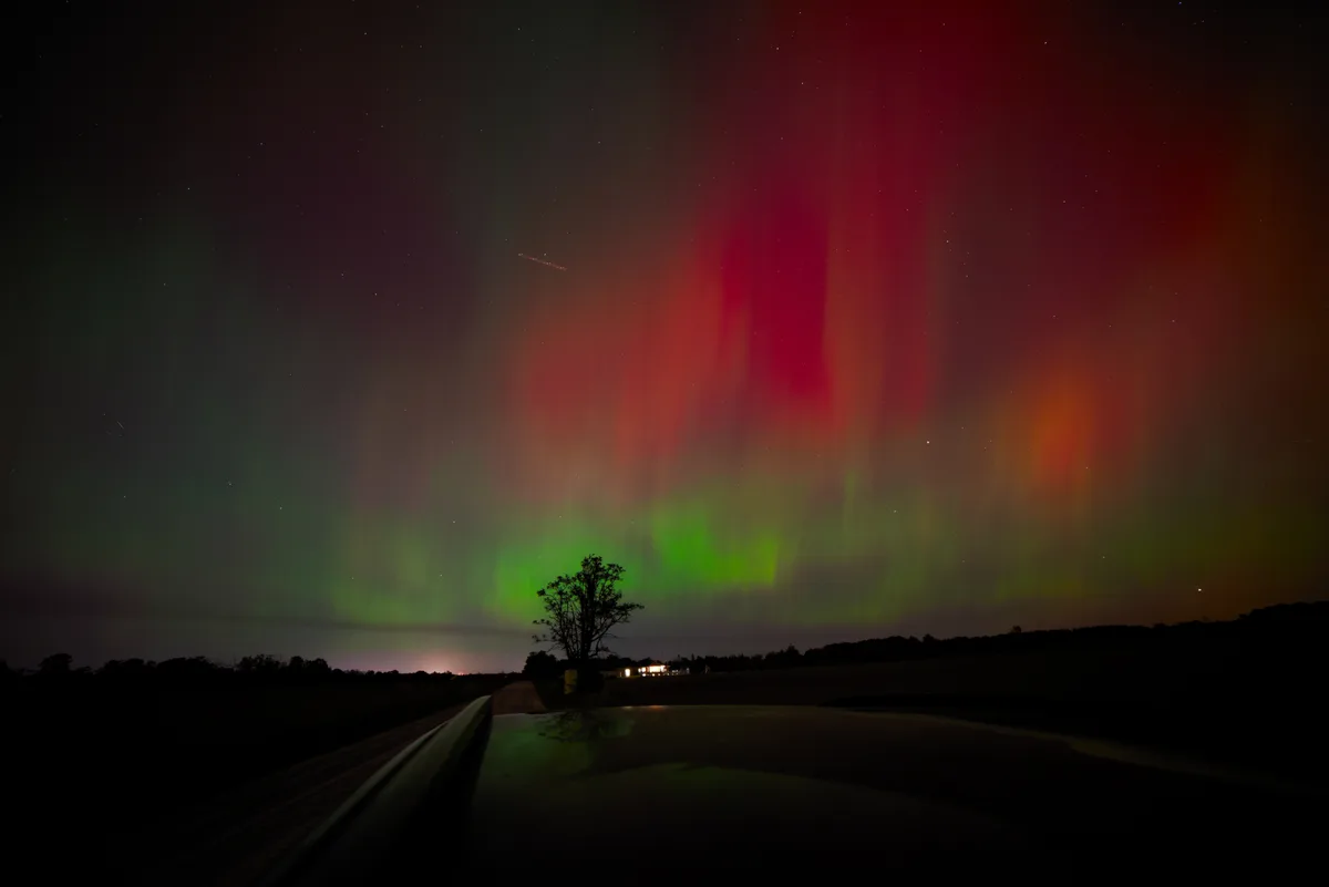 A nighttime photo taken from inside a car shows a vibrant aurora borealis with red and green curtains across a starry sky. A…