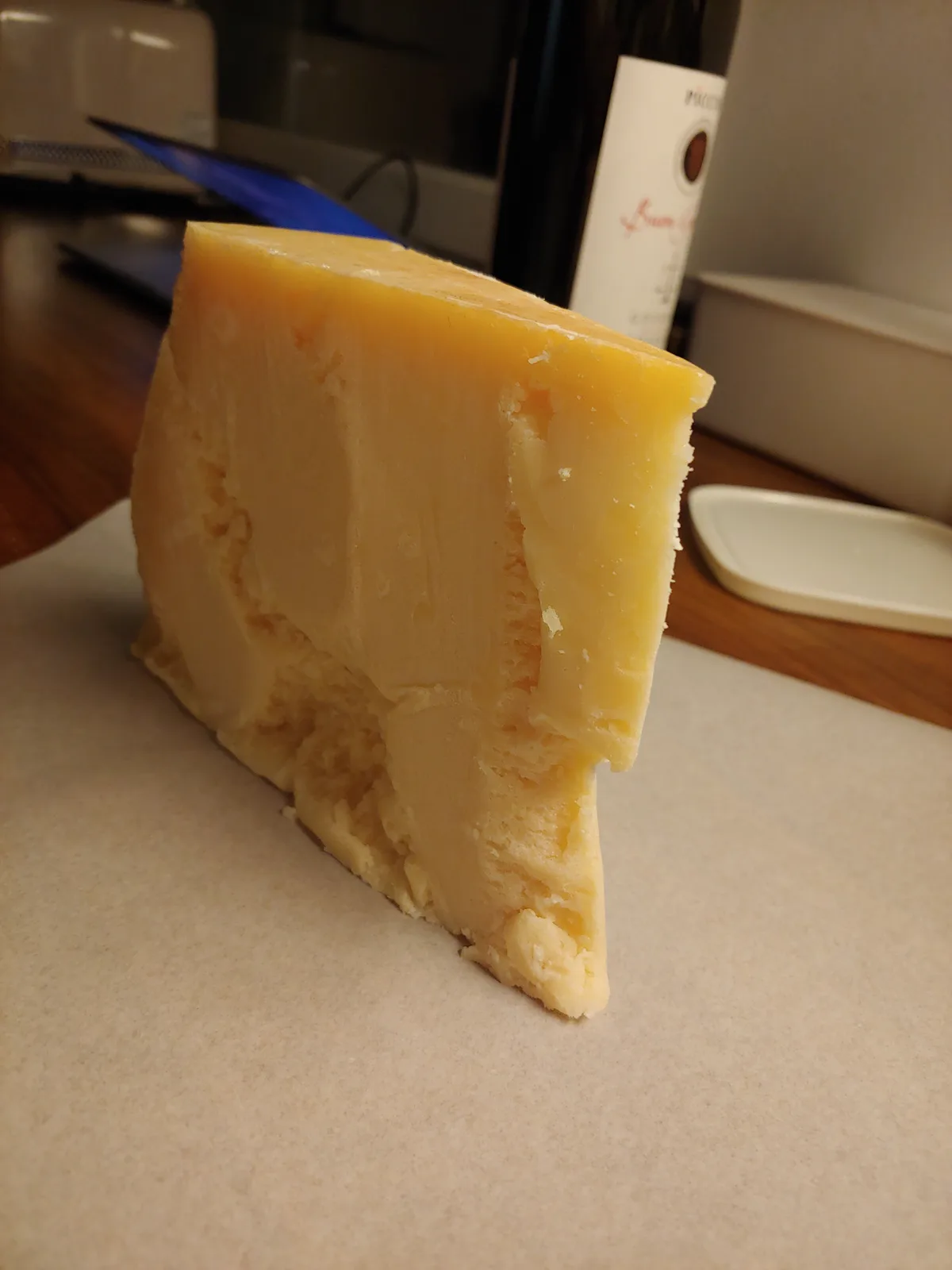A large, wedge-shaped slice of yellow cheese stands upright on parchment paper, with a wine bottle and laptop visible in the…