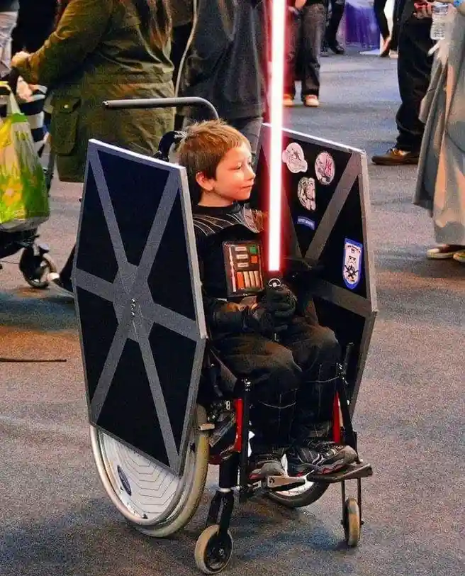 A young boy in a wheelchair is dressed as a Star Wars character, holding a red lightsaber and flanked by two large, black…