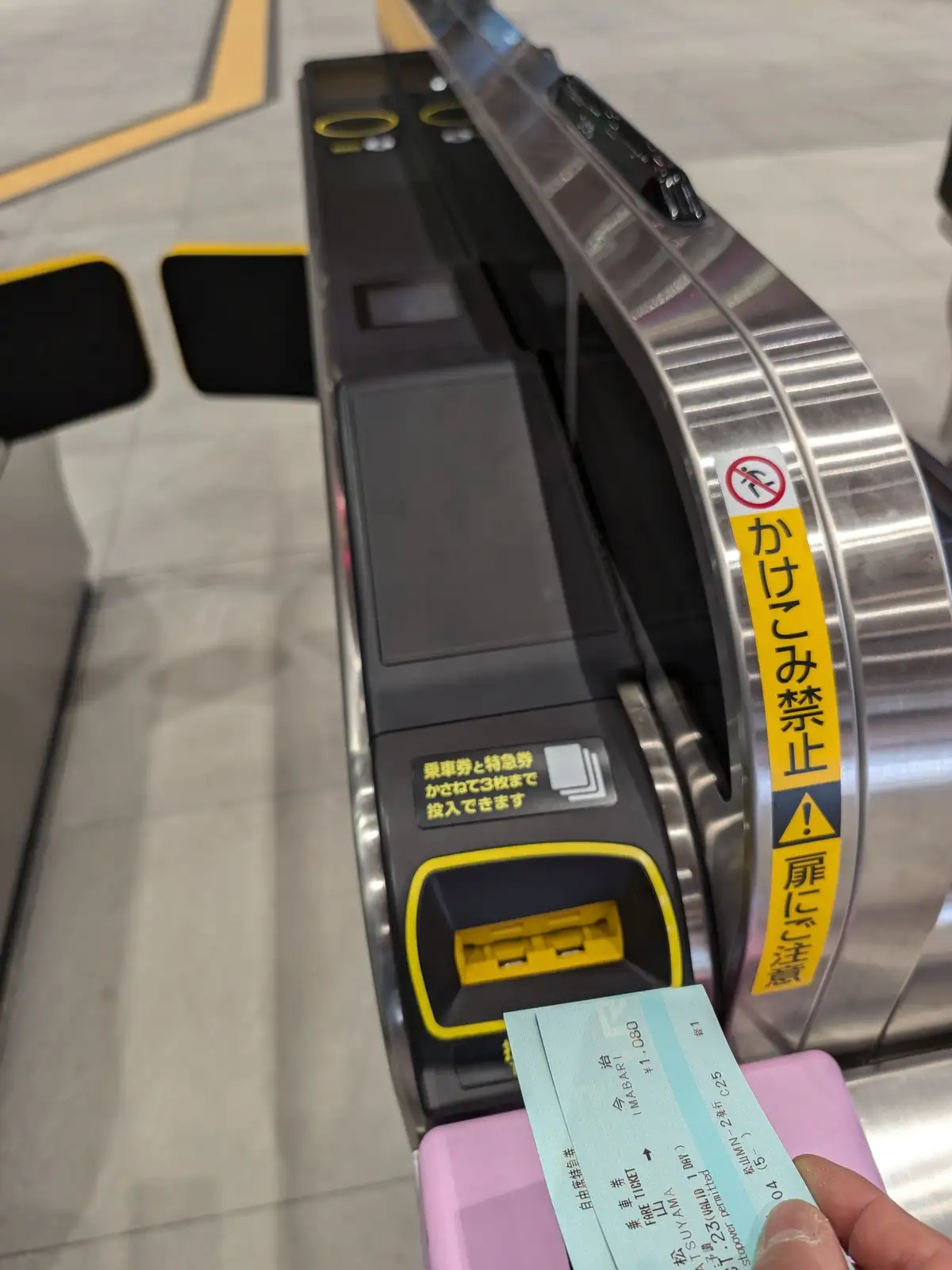 A close-up photo shows a hand holding a light blue Japanese train ticket in front of a ticket validation machine at a…