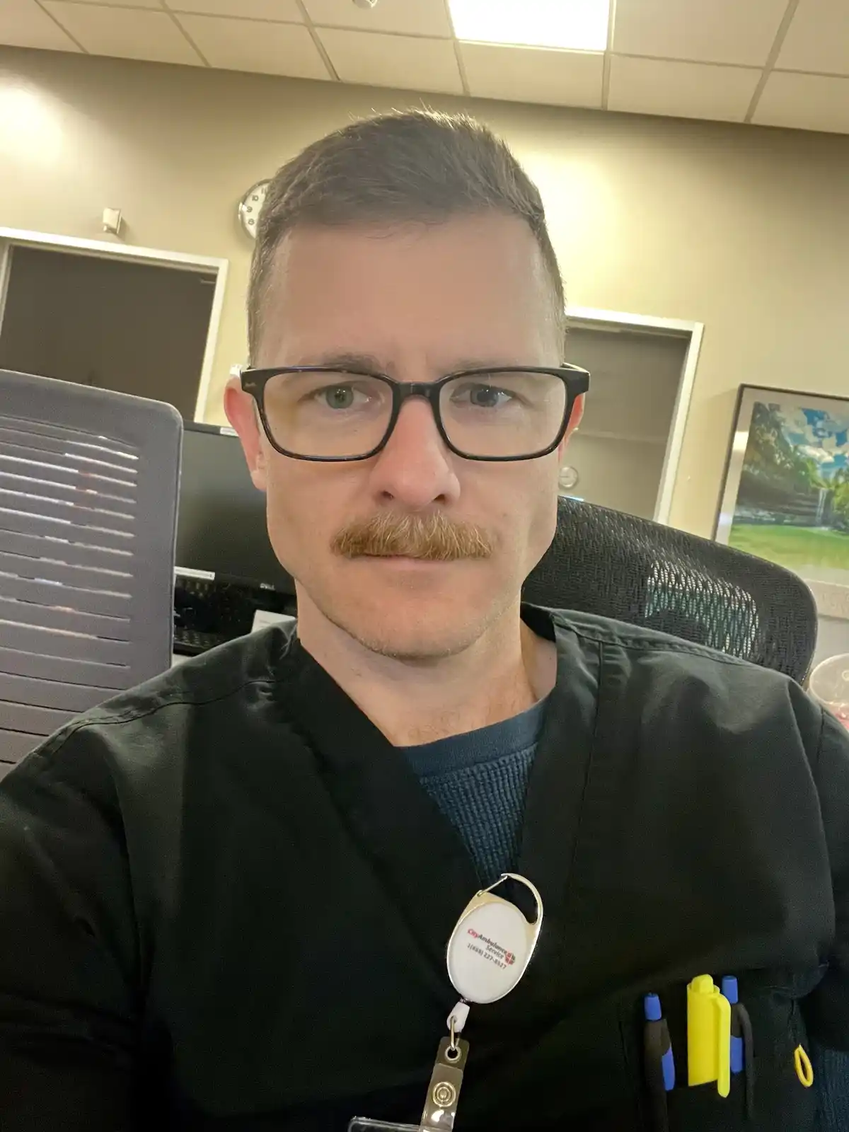 A man in black scrubs and glasses, with a mustache, sits in an office chair. He wears a lanyard with a badge that reads…