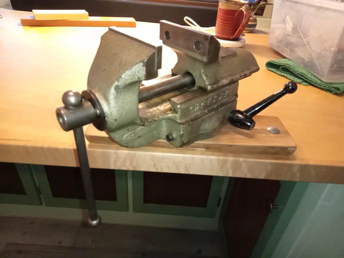 A green metal bench vise is mounted on a wooden board atop a kitchen counter, with its jaws open and handle visible. In the…