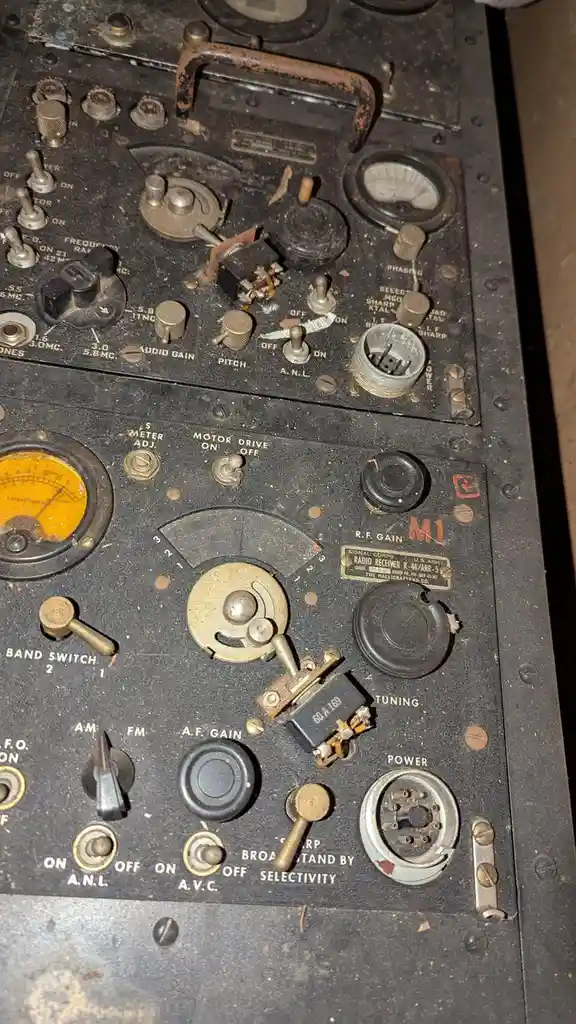 A close-up photo of a very old, dusty, and worn military radio receiver with many knobs, dials, and switches labeled with…