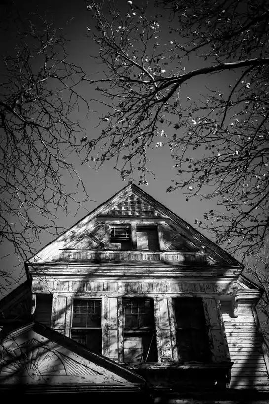 A black-and-white photograph shows the upper portion of a dilapidated, weathered wooden house with peeling paint and broken…