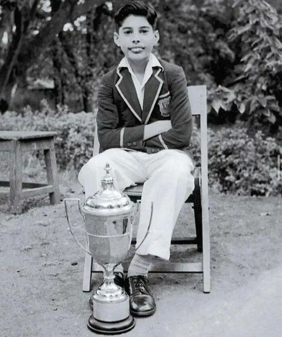 A black-and-white photo shows a young boy in a school uniform sitting on a wooden chair outdoors with a large silver trophy…