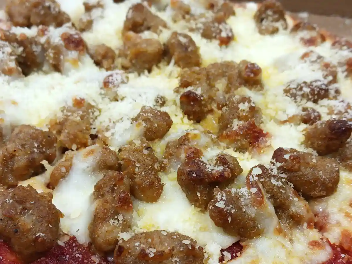 A close-up photo shows a pizza with a visible layer of red sauce, melted white cheese, and numerous browned sausage crumbles…