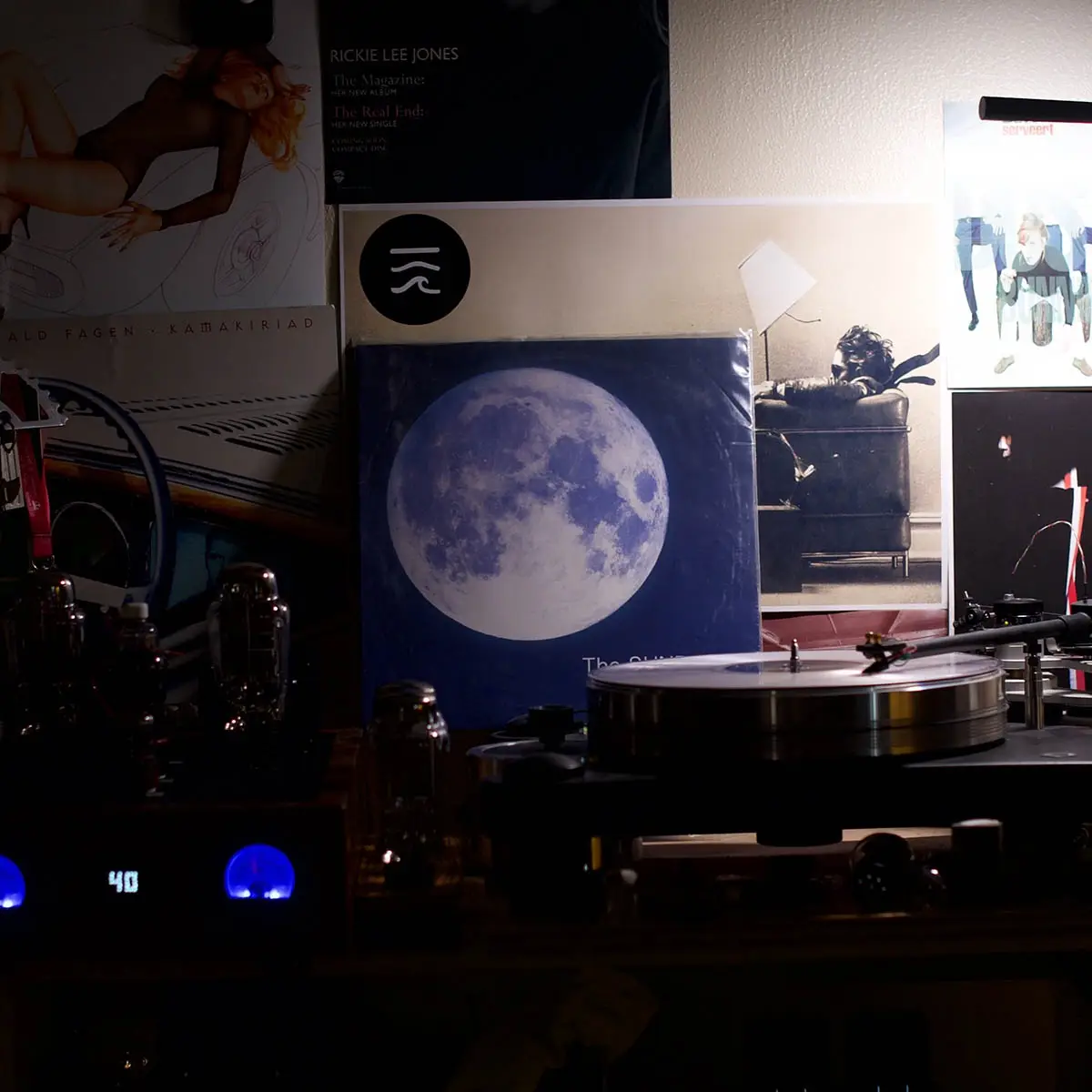 A dimly lit room features a turntable with a record on it, surrounded by various vinyl record album covers on the wall…