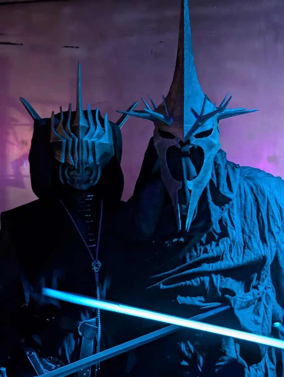 Two figures dressed as Sauron and a Nazgûl from The Lord of the Rings stand side by side in dark, hooded robes under blue…