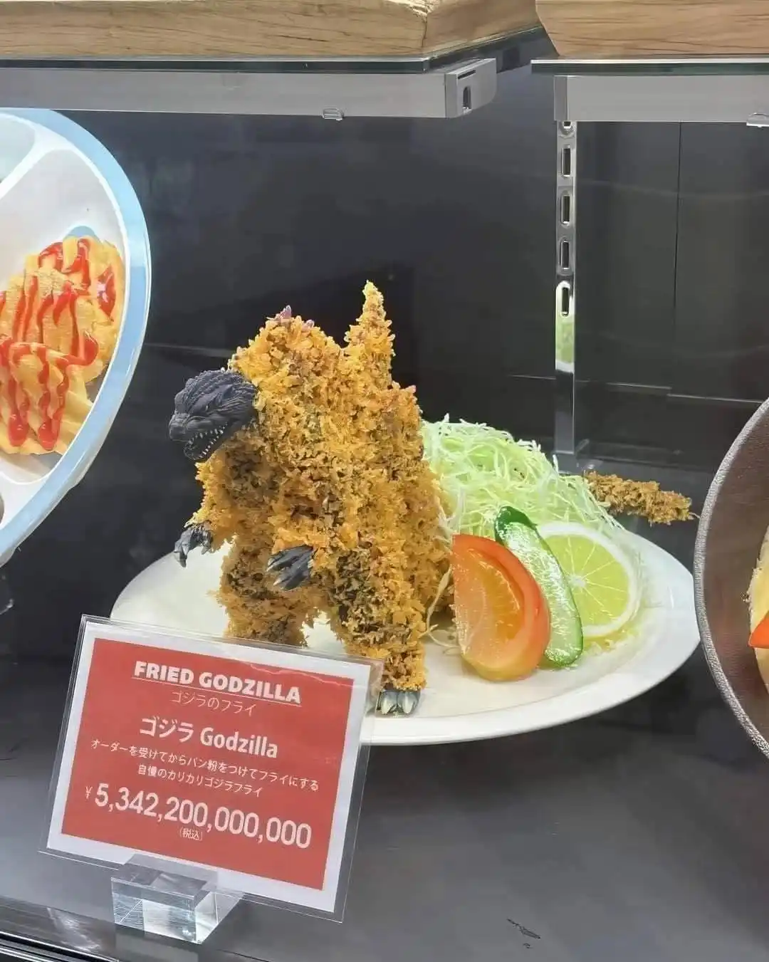 A plate of fried Godzilla-shaped food, covered in breadcrumbs, is displayed in a glass case with a red sign. The sign reads…