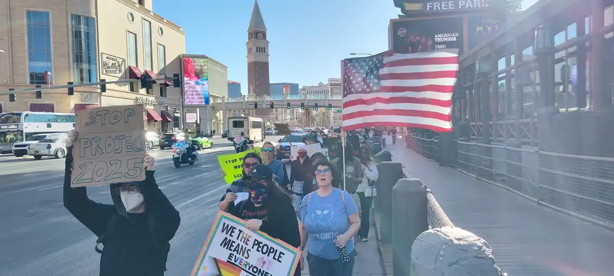 A group of protesters marches on a Las Vegas street, holding signs including 'STOP PROJECT 2025' and 'WE THE PEOPLE MEANS…
