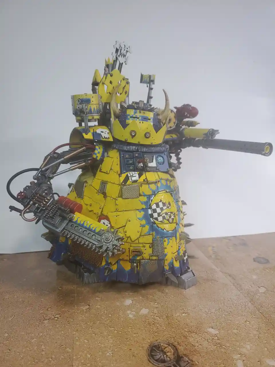 A heavily customized, yellow and blue painted Warhammer 40k tank model with multiple weapons, horns, and a crown, standing…