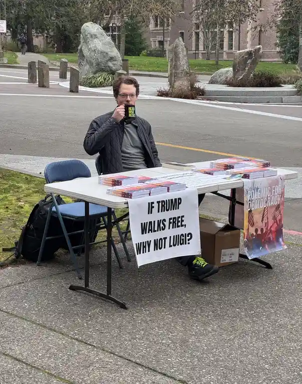 A man sits at a folding table outdoors, sipping from a mug that reads 'READ RISE RESIST', surrounded by stacks of books and…