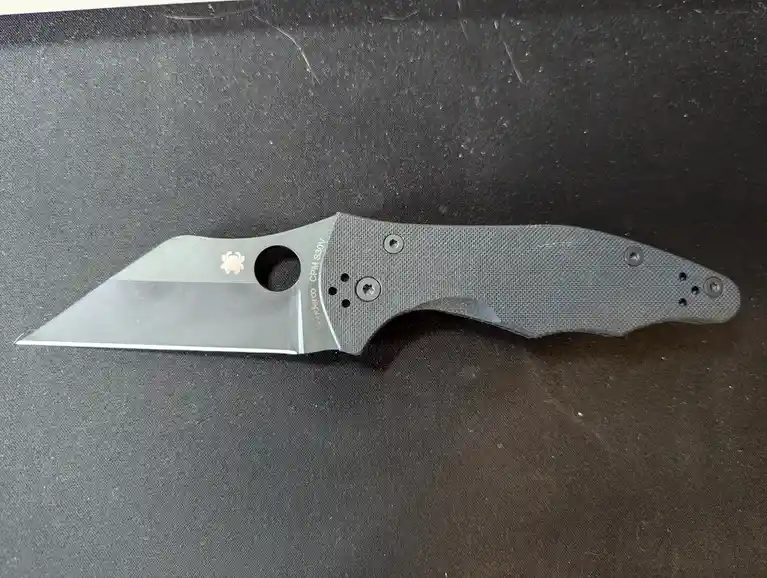 A black Spyderco folding knife with a textured handle and a circular hole near the blade's base lies on a dark, textured…
