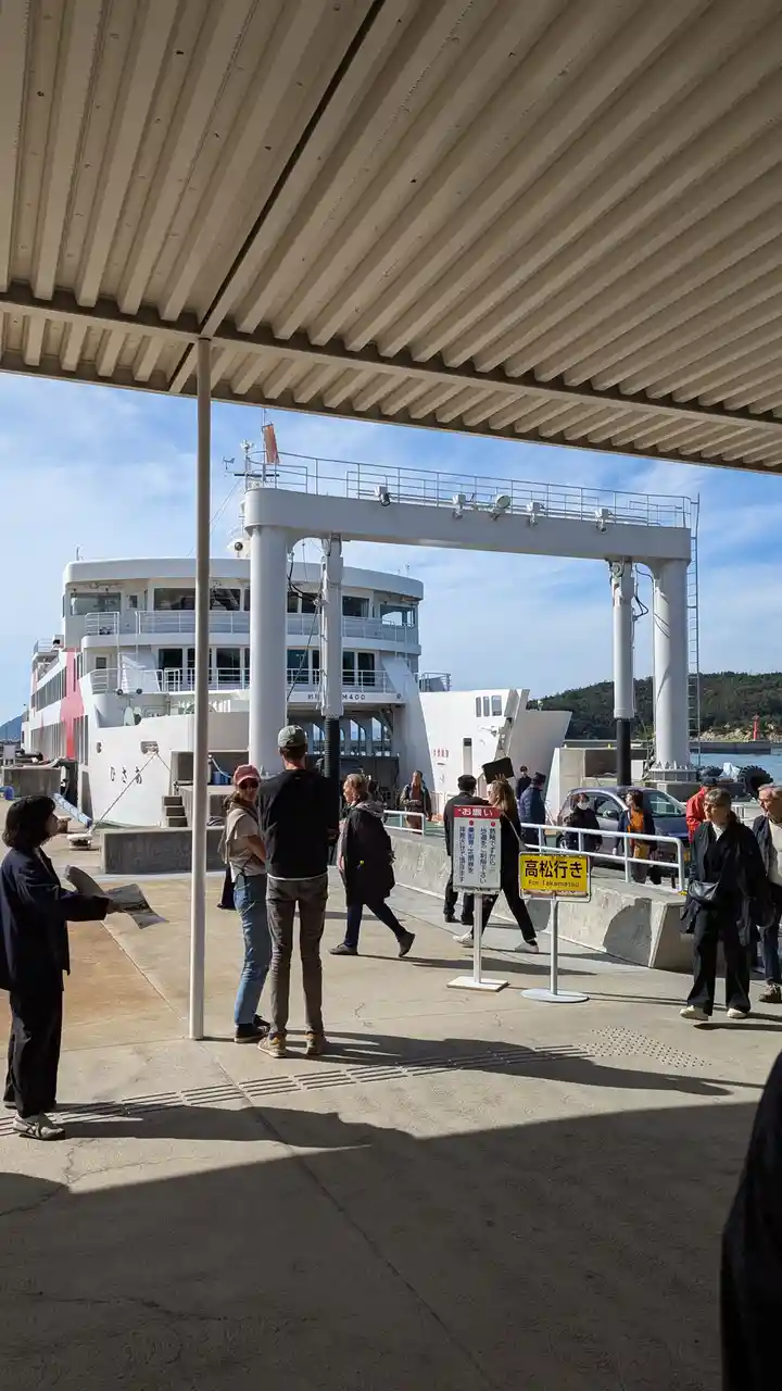 A photo of a ferry dock with people waiting, featuring a yellow sign that reads '高松行き For Takamatsu' in both Japanese and…