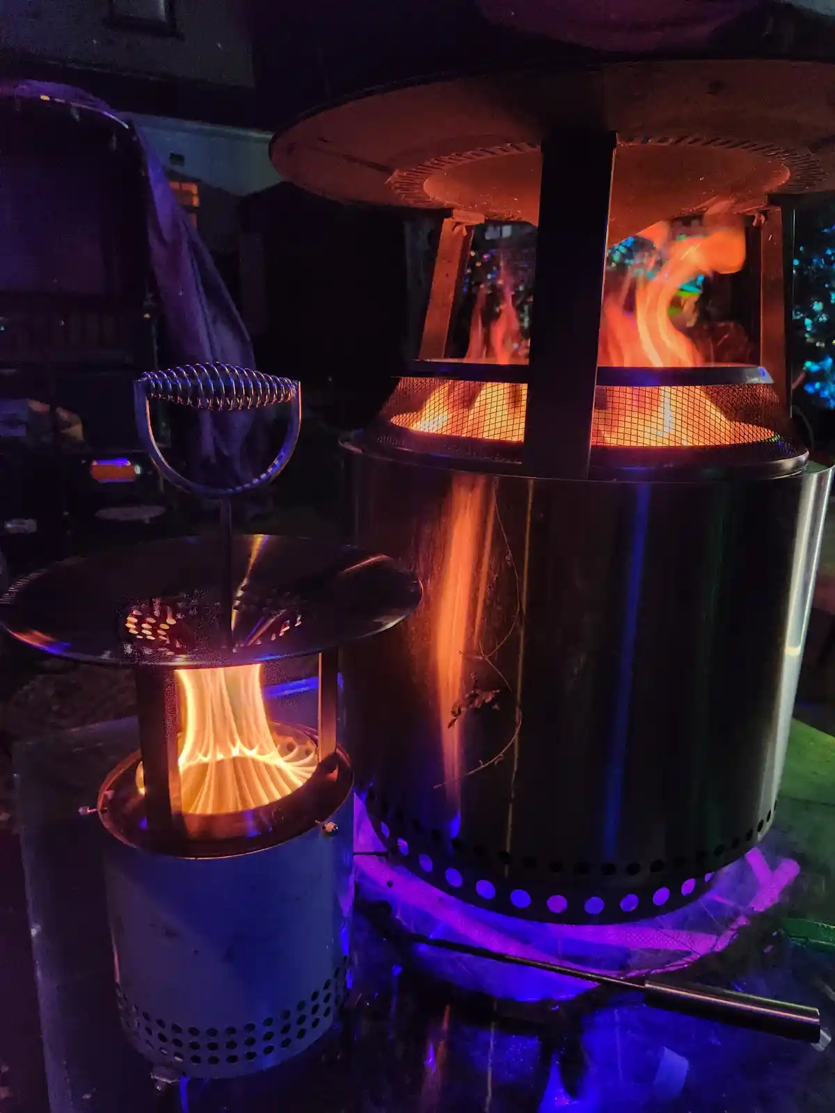 Two outdoor fire pits are lit up at night, one small and one large, both glowing with orange flames. The smaller one has a…