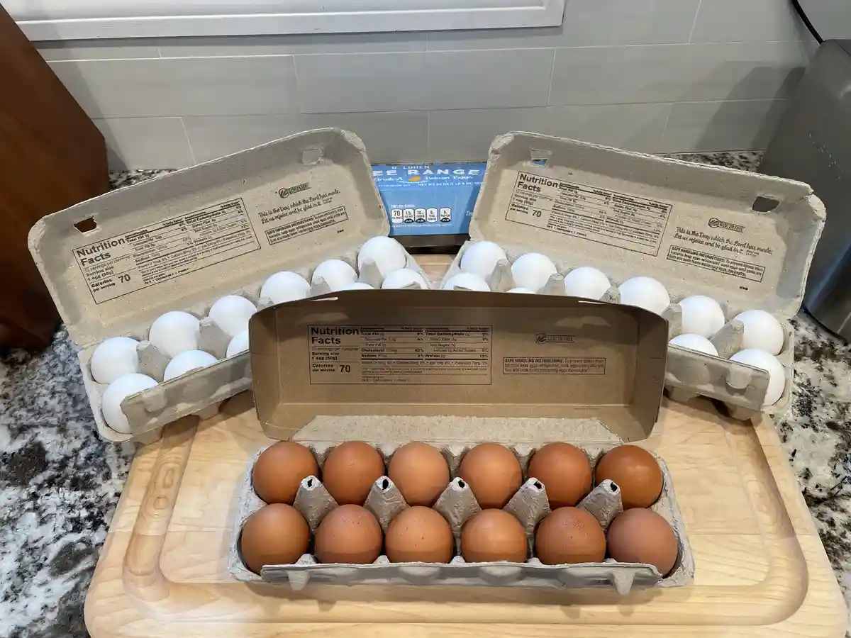 Three open cardboard egg cartons sit on a wooden cutting board on a kitchen counter. The front carton holds brown eggs…